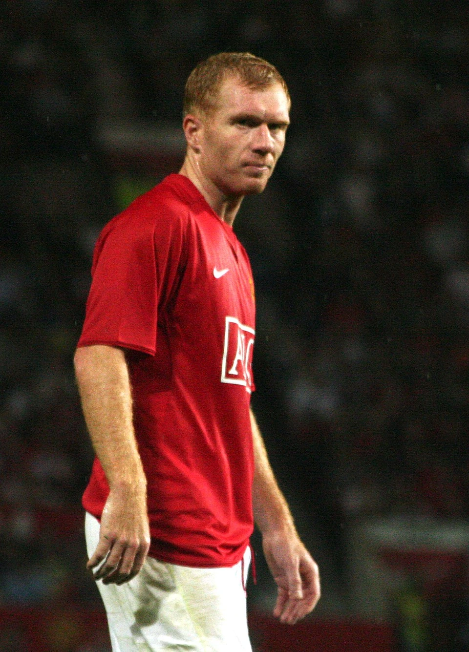 Paul Scholes in a Manchester United red jersey during a football match, looking over his shoulder against a dark stadium