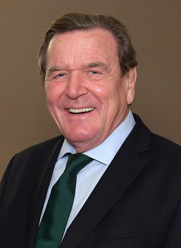 Gerhard Schröder smiling in a suit and green tie against a plain background.