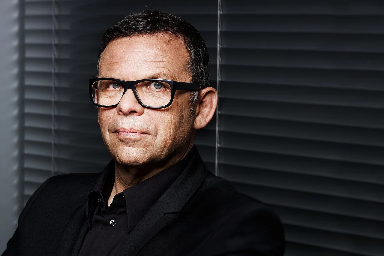 Portrait photo of Peter Schreyer wearing glasses and a black suit against dark blinds