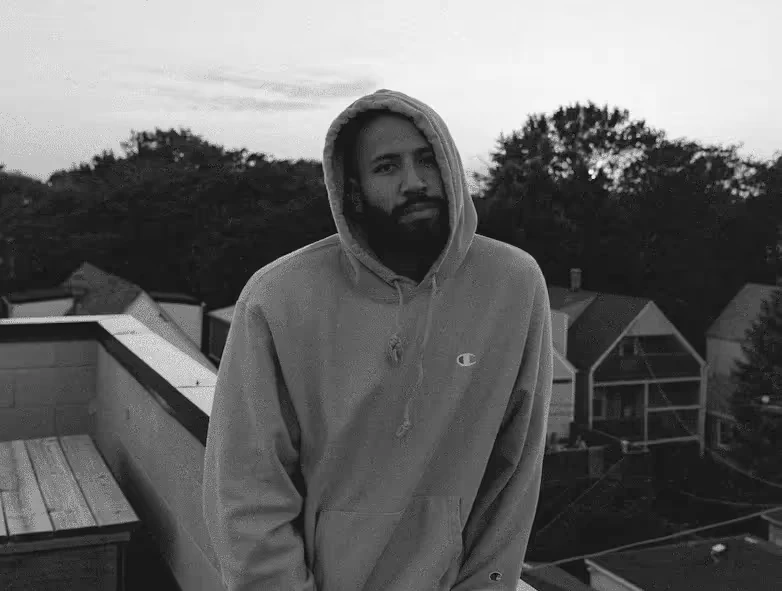 Black-and-white photo of Cole Schafer standing outdoors in a hoodie on a rooftop, with houses and trees in the backgroun